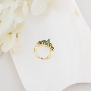 May include: A gold-colored nose ring with a row of green gemstones. The ring is set against a white background with white flowers in the upper left corner. The gemstones are a mix of light and dark green.
