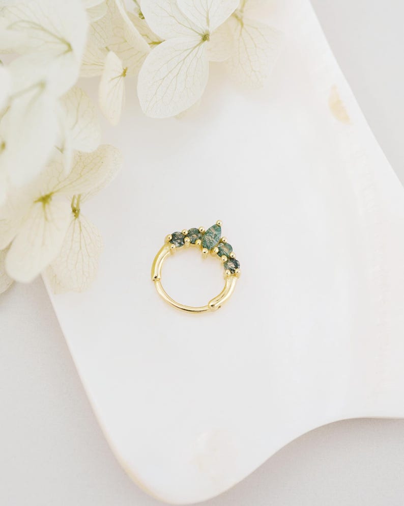 May include: A gold-colored nose ring with a row of green gemstones. The ring is set against a white background with white flowers in the upper left corner. The gemstones are a mix of light and dark green.