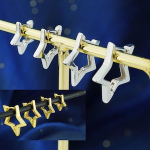 May include: Silver star-shaped hoop earrings displayed on a gold bar. The earrings are open-hoop style, with a star design. A second image shows gold star earrings on a black bar. The background is a gradient of blue.