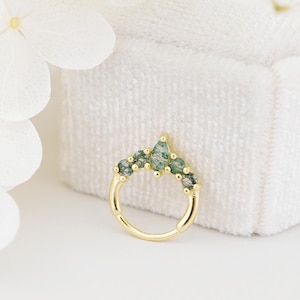 May include: A gold-colored nose ring with a series of green gemstones. The gemstones are arranged in a curved pattern, with a larger, teardrop-shaped stone at the top center. The ring is set against a white background.