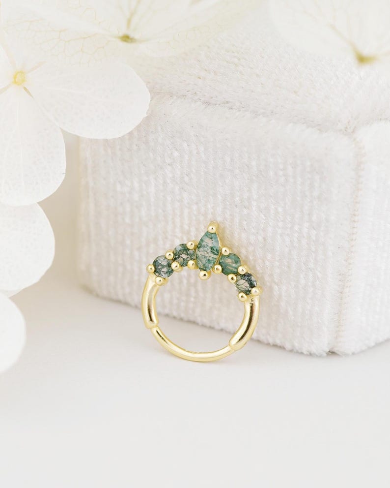 May include: A gold-colored nose ring with a series of green gemstones. The gemstones are arranged in a curved pattern, with a larger, teardrop-shaped stone at the top center. The ring is set against a white background.