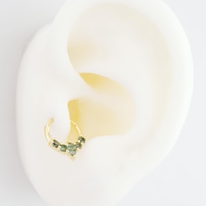 May include: A gold-colored hoop earring with a cluster of green gemstones. The earring is attached to a white ear model, showcasing the jewelry's design and color contrast. The gemstones are arranged in a decorative pattern.