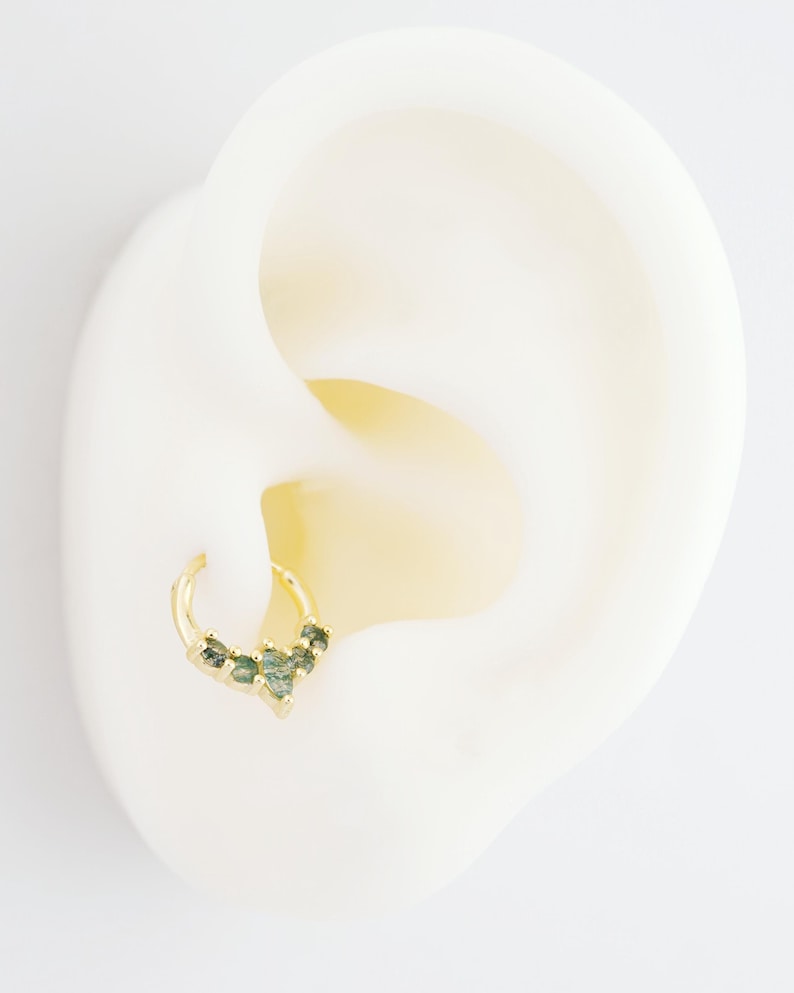 May include: A gold-colored hoop earring with a cluster of green gemstones. The earring is attached to a white ear model, showcasing the jewelry's design and color contrast. The gemstones are arranged in a decorative pattern.