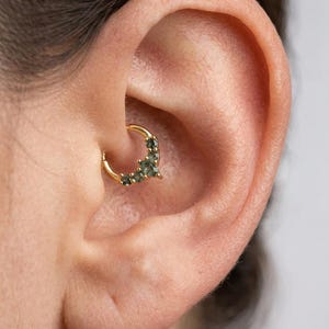 May include: A gold-colored septum ring with small, dark green gemstones. The jewelry is set in a curved design and is worn in a person's ear. The ear is a light skin tone.