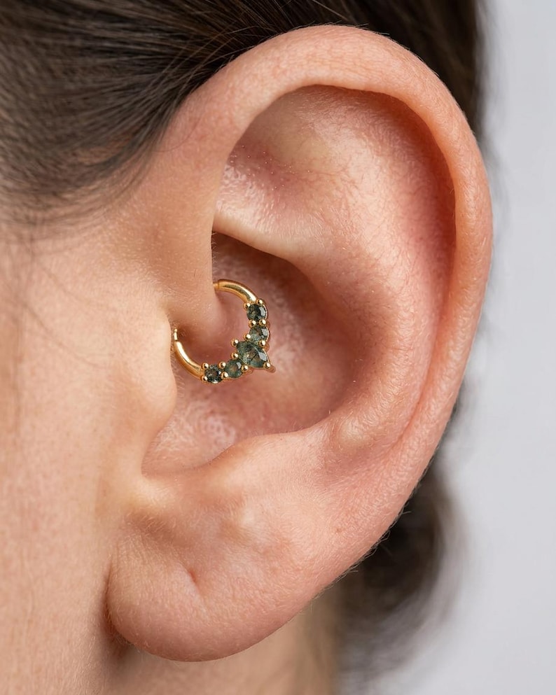 May include: A gold-colored septum ring with small, dark green gemstones. The jewelry is set in a curved design and is worn in a person's ear. The ear is a light skin tone.