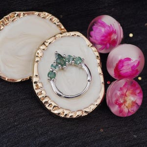 May include: A silver ring with five green gemstones is displayed on a white and gold-edged decorative tray. Three clear orbs with pink flowers inside are also present, set against a dark background.