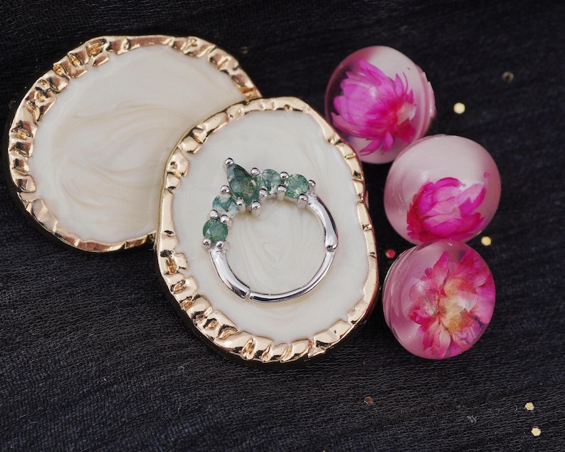 May include: A silver ring with five green gemstones is displayed on a white and gold-edged decorative tray. Three clear orbs with pink flowers inside are also present, set against a dark background.