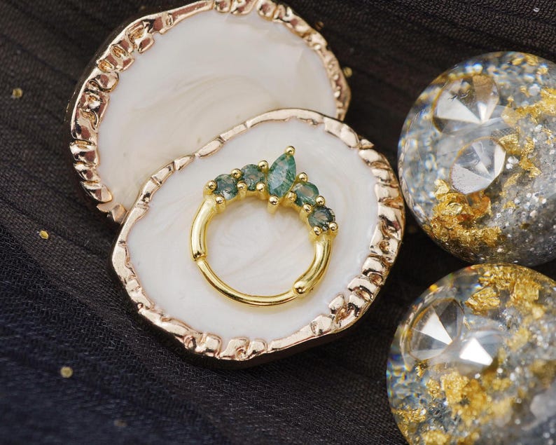 May include: A gold-colored ring with a row of five teal-colored gemstones, set on a white and gold-edged display. Two decorative spheres with gold flakes and clear gems are also visible.