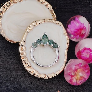 May include: A silver ring with a cluster of green gemstones, displayed on a white and gold decorative tray. The ring is surrounded by three clear resin spheres, each containing a pink flower. The background is a dark fabric.