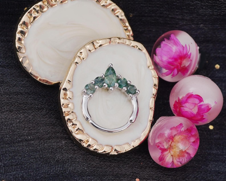 May include: A silver ring with a cluster of green gemstones, displayed on a white and gold decorative tray. The ring is surrounded by three clear resin spheres, each containing a pink flower. The background is a dark fabric.
