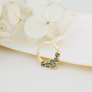 May include: A gold-colored septum ring with a cluster of small, green gemstones. The ring is circular and has a hinged closure. The gemstones are a mix of light and dark green, and are set in a delicate, decorative pattern.