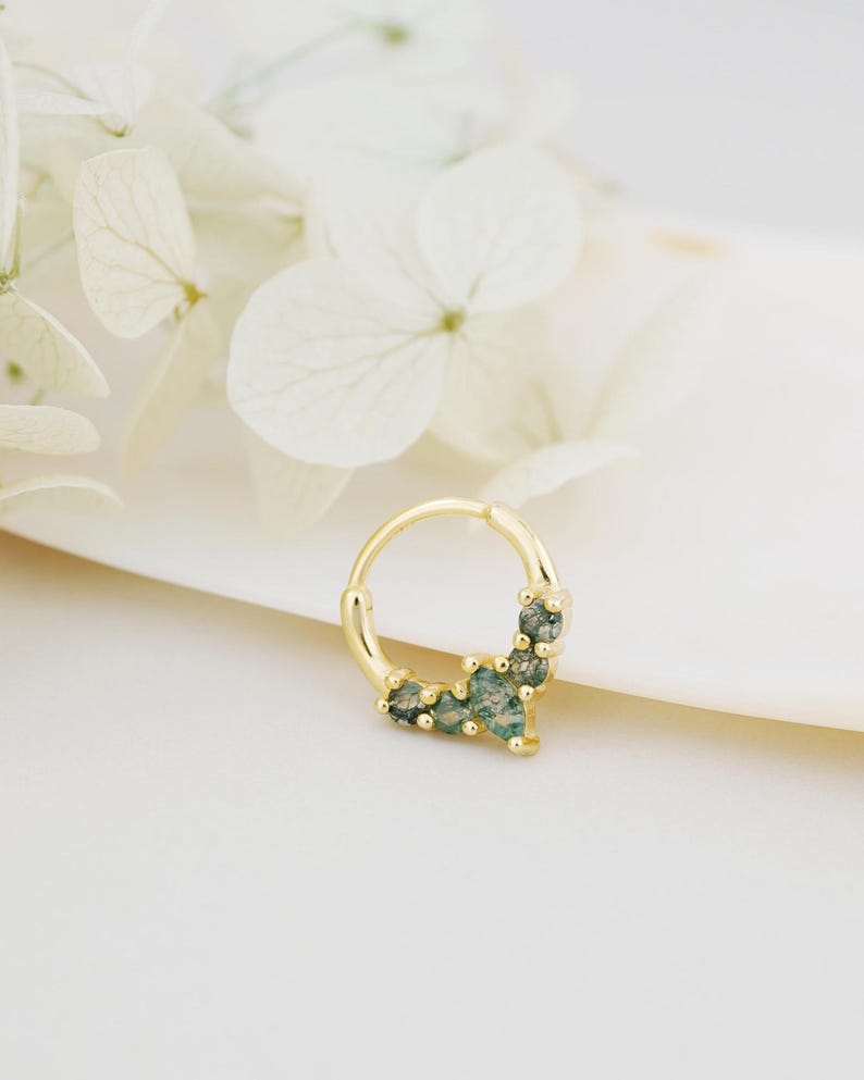 May include: A gold-colored septum ring with a cluster of small, green gemstones. The ring is circular and has a hinged closure. The gemstones are a mix of light and dark green, and are set in a delicate, decorative pattern.