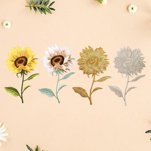 May include: Four embroidered sunflower patches arranged in a row. The first is yellow with a brown centre, the second is white with a brown centre, the third is metallic gold, and the fourth is metallic silver. Each features a stem and leaves.