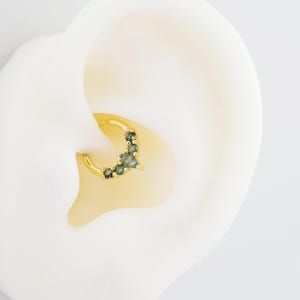 May include: A gold-colored hoop earring with a cluster of small, green gemstones. The earring is set against a white background, highlighting the jewelry's design and color. The gemstones are a mix of light and dark green.