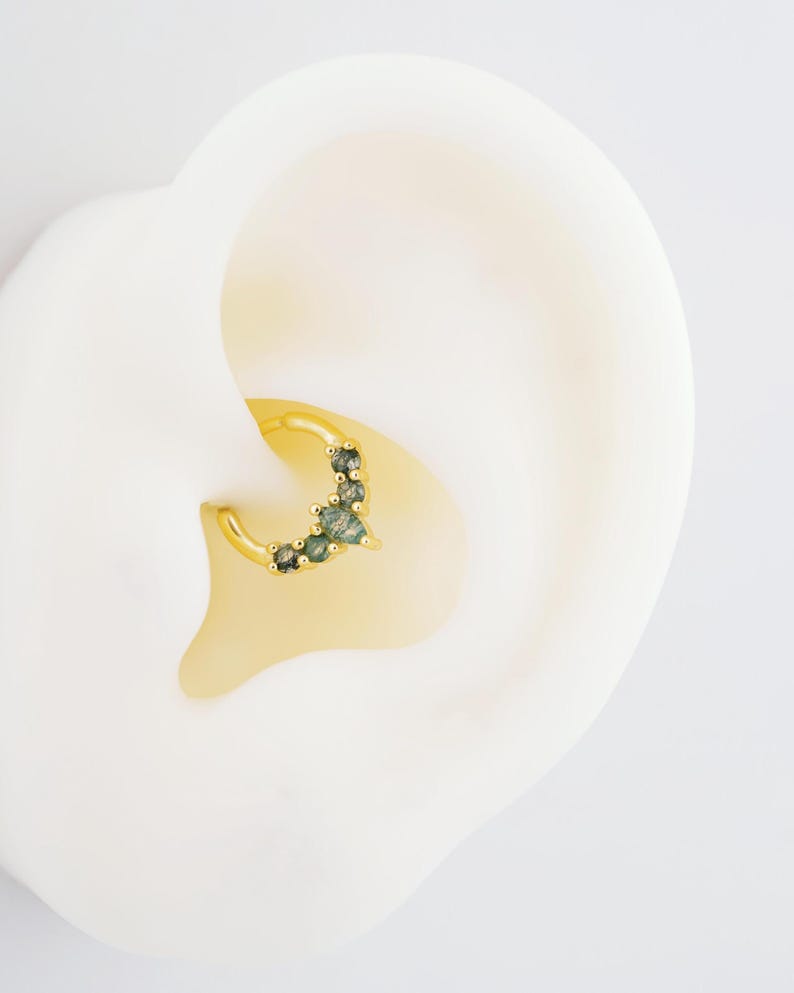 May include: A gold-colored hoop earring with a cluster of small, green gemstones. The earring is set against a white background, highlighting the jewelry's design and color. The gemstones are a mix of light and dark green.
