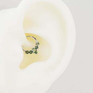 May include: A silver septum ring featuring a cluster of green gemstones. The ring is designed with a hinged closure and is shown in a close-up view, highlighting the details of the jewelry.