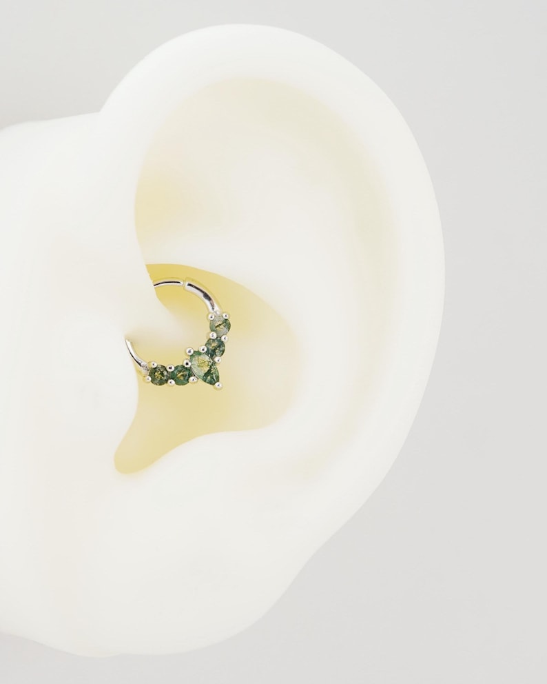 May include: A silver septum ring featuring a cluster of green gemstones. The ring is designed with a hinged closure and is shown in a close-up view, highlighting the details of the jewelry.
