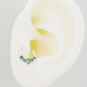 May include: A silver hoop earring with five green gemstones arranged in a V-shape. The earring is shown on a white ear model, highlighting the jewelry's design and placement. The gemstones have a subtle sparkle, adding a touch of elegance.