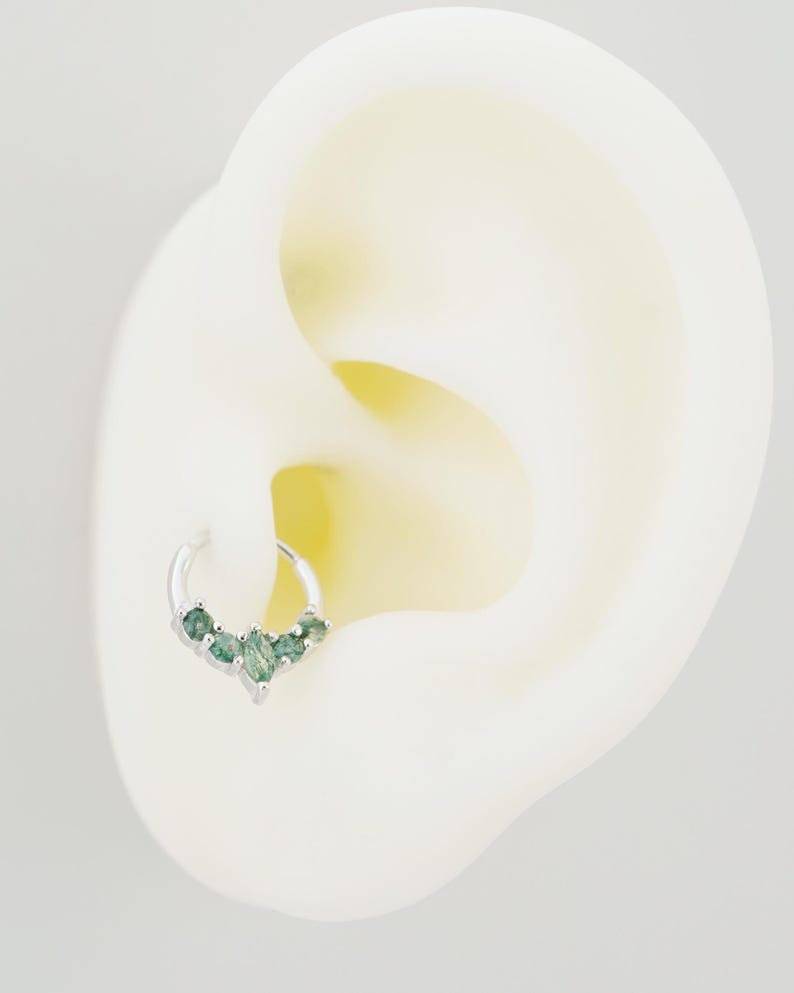 May include: A silver hoop earring with five green gemstones arranged in a V-shape. The earring is shown on a white ear model, highlighting the jewelry's design and placement. The gemstones have a subtle sparkle, adding a touch of elegance.