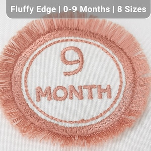 Puede incluir: Un parche bordado redondo, color melocotón, con un borde difuso. El parche presenta el número "9" y la palabra "MONTH" en un color melocotón a juego, sobre un fondo blanco. El texto "Fluffy Edge | 0-9 Months | 8 Sizes" está en la parte superior.