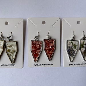 May include: Three pairs of dangle earrings with different designs. The first pair features dried flowers in a clear resin, the second pair has a copper glitter design, and the third pair has dried flowers in a clear resin with a silver frame. Each pair is displayed on a white card with the text "EVAS ART AND SENSORY".