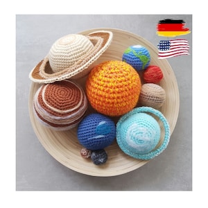Puede incluir: Un conjunto de planetas de ganchillo, que incluye una Tierra azul y blanca, un Marte rojo, un Júpiter marrón, un Sol amarillo y un Neptuno azul. Los planetas están dispuestos en una bandeja de madera.