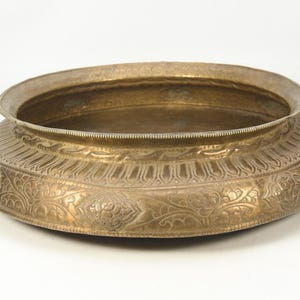May include: A low, round, antique brass bowl with an ornate, embossed floral pattern. The bowl has a wide, shallow basin and a slightly raised rim. The exterior is decorated with detailed relief designs.