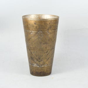 May include: A tall, brass drinking glass with an intricate, engraved pattern. The glass has a conical shape and a smooth, polished finish.