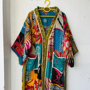 Könnte beinhalten: Kimono-ähnlicher Bademantel im Patchwork-Stil mit V-Ausschnitt und Taschen. Der Bademantel weist eine Vielzahl farbenfroher Stoffflicken in Rot-, Blau-, Gelb- und Grüntönen auf. Der Bademantel hängt an einem gelben Kleiderbügel.