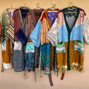 May include: A collection of six colorful, patchwork kimonos hanging on a wall. Each kimono features a unique combination of patterns and colors, including shades of green, blue, brown, and gold. The kimonos are made of a silky material and have a relaxed, open design.