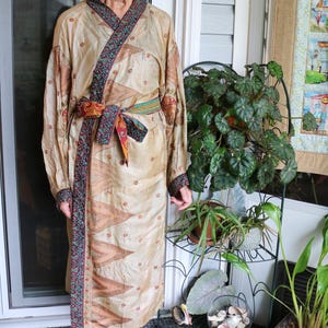 May include: A long-sleeved robe in a light beige hue featuring a repeating pattern of small red floral motifs. The garment includes a dark trim with a multi-colored paisley design along the collar, cuffs, and a matching tie-waist belt.