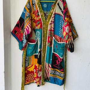 Puede incluir: Una colorida bata tipo kimono de patchwork con cuello en V y bolsillos. La bata presenta una variedad de patrones de tela en tonos de azul, rojo, amarillo y verde. La bata está colgada en una percha amarilla contra una pared blanca.