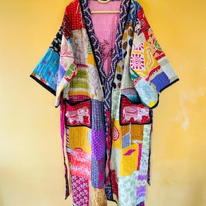 May include: A patchwork kimono robe with a variety of colorful fabric squares. The robe has a pink interior and black trim. The sleeves are wide and the robe is hanging on a yellow wall.