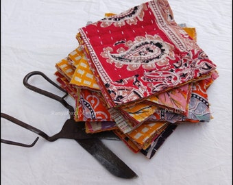Könnte beinhalten: Ein Stapel bunter, gemusterter Stoffquadrate in Rot-, Gelb- und Orangetönen. Der Stoff weist Paisley- und Blumenmuster auf. Eine antike Metallschere liegt auf der weißen Oberfläche neben dem Stoff.