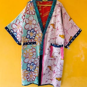 Peut inclure: Robe de style kimono en patchwork avec un motif floral et d'oiseaux. La robe présente un mélange de tissus bleu clair, rose et rouge, avec des bordures et des poches bleu foncé. La robe est suspendue à un cintre jaune sur fond jaune.