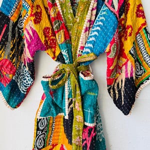 Op de afbeelding: Een kleurrijke patchwork badjas met een strikceintuur. De badjas heeft verschillende stofpatronen in blauw, geel, rood, groen en roze. De badjas is gemaakt van gewatteerde stof en heeft wijde mouwen.