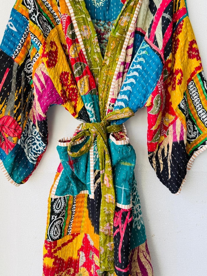 Op de afbeelding: Een kleurrijke patchwork badjas met een strikceintuur. De badjas heeft verschillende stofpatronen in blauw, geel, rood, groen en roze. De badjas is gemaakt van gewatteerde stof en heeft wijde mouwen.