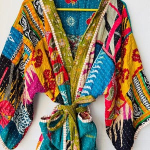Op de afbeelding: Een kleurrijke patchwork badjas met een strikceintuur. De badjas heeft verschillende stofpatronen in blauw, geel, groen, roze en zwart. De badjas hangt aan een gele hanger.