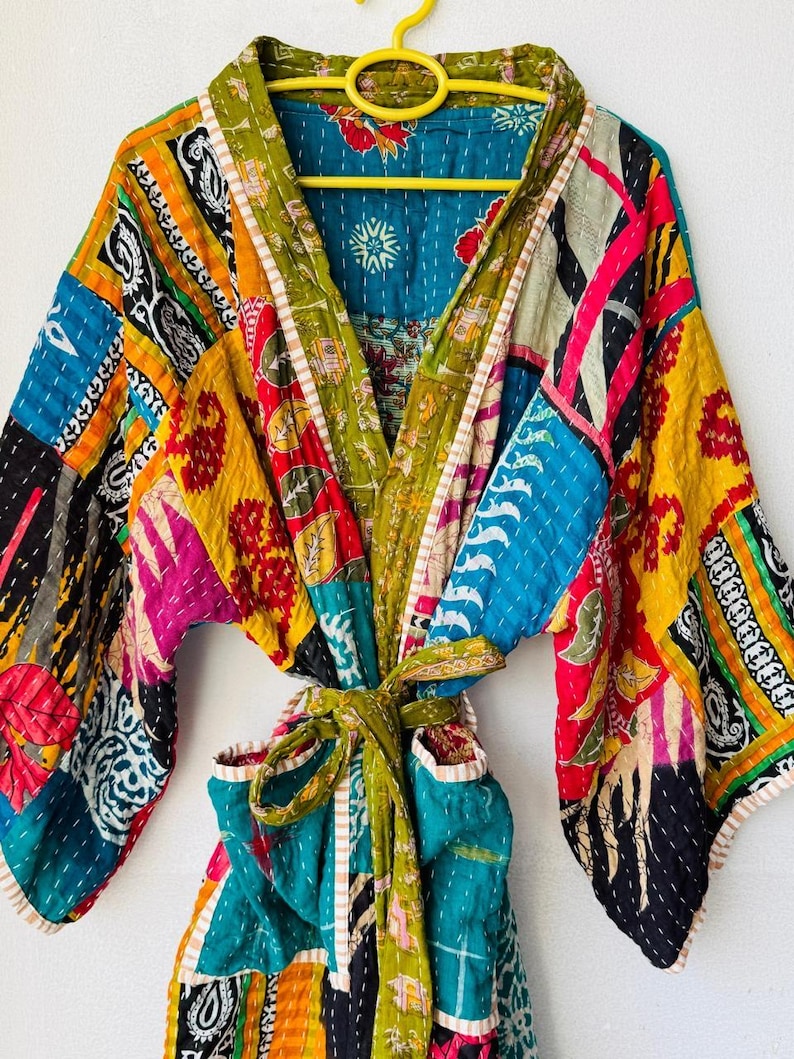 Op de afbeelding: Een kleurrijke patchwork badjas met een strikceintuur. De badjas heeft verschillende stofpatronen in blauw, geel, groen, roze en zwart. De badjas hangt aan een gele hanger.