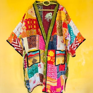 Könnte beinhalten: Eine farbenfrohe Patchwork-Kimono-Jacke mit einer Vielzahl von Stoffmustern und -farben, darunter Rot, Gelb, Rosa und Grün. Die Jacke hat eine grüne Einfassung und zwei Taschen. Sie hängt an einer gelben Wand.