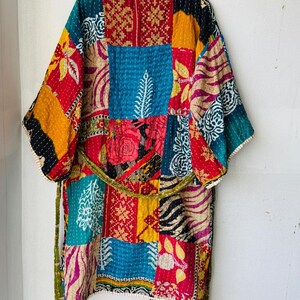 Op de afbeelding: Een patchwork kimono-achtige badjas met een riem. De badjas heeft een verscheidenheid aan stoffen vierkanten met patronen in rood, blauw, geel en groen. De badjas hangt aan een gele hanger tegen een witte muur.