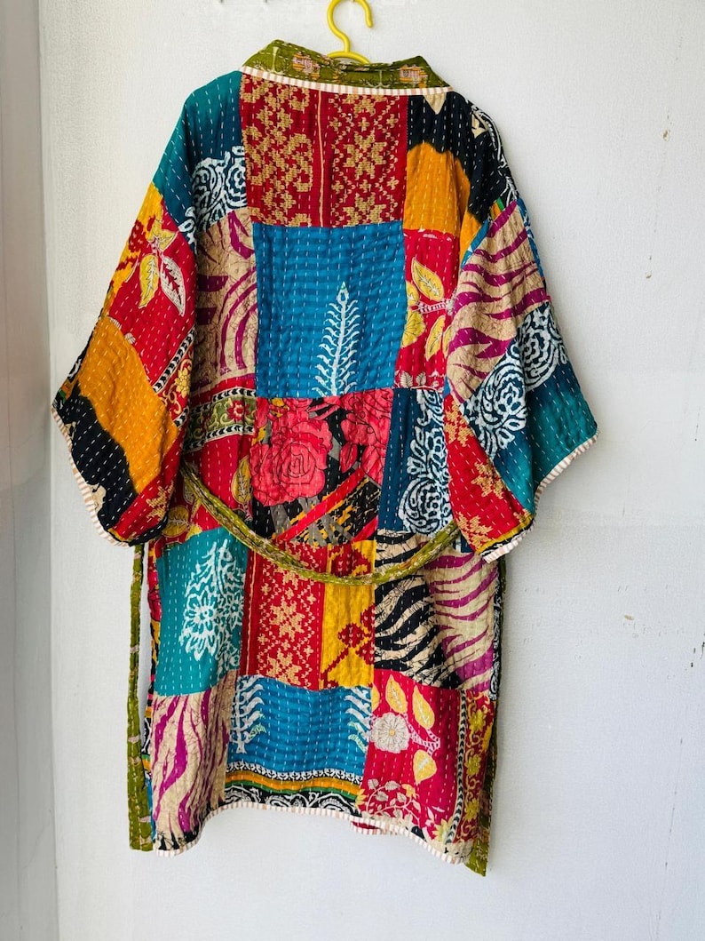 Op de afbeelding: Een patchwork kimono-achtige badjas met een riem. De badjas heeft een verscheidenheid aan stoffen vierkanten met patronen in rood, blauw, geel en groen. De badjas hangt aan een gele hanger tegen een witte muur.
