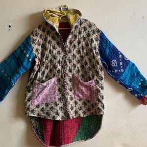 May include: A hooded jacket featuring a beige base with a black floral pattern, long sleeves with blue and white tie-dye style sections, two pink front pockets, and a curved hemline displaying red and green fabric panels with visible stitching.