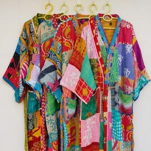 Può includere: Cinque kimono patchwork colorati sono appesi a grucce gialle. I kimono presentano una varietà di motivi e colori, tra cui rosso, blu, verde e rosa. Ogni kimono ha un design unico.