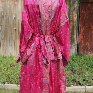May include: A long-sleeved, floor-length robe in a vibrant magenta pink hue, featuring a repeating gold paisley pattern throughout. The garment includes a matching fabric tie belt at the waist and a wide, contrasting shawl collar with intricate detailing.