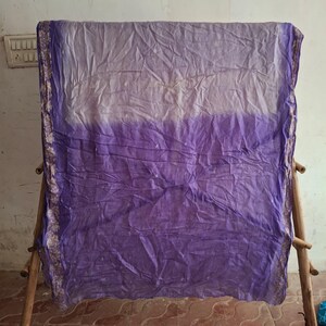 May include: A rectangular fabric scarf featuring an ombre design that transitions from a light lavender at the top to a deep violet at the bottom. The edges are finished with a decorative border in a muted gold tone with intricate floral patterns.