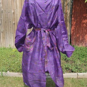 May include: A long-sleeved purple robe featuring a subtle, intricate pattern in shades of magenta and gold. The garment includes a wide shawl collar, a matching tie belt at the waist, and two front pockets, displayed on a black metal stand.