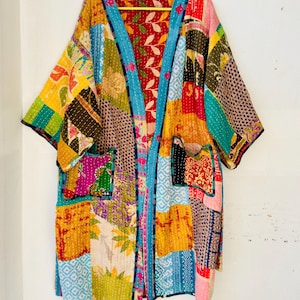 Op de afbeelding: Een patchwork kimono-achtige robe met een levendige mix van kleuren en patronen. De robe heeft een turkooizen rand, twee voorzakken en een bloemenvoering. Het ontwerp bevat stoffen in tinten geel, roze, groen en blauw.