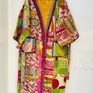 May include: A patchwork kimono robe with a vibrant mix of colors and patterns. The robe features a yellow lining, green pockets, and a variety of fabric squares with floral and paisley designs. The robe is hanging on a yellow hanger.