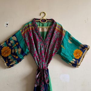 May include: A kimono-style robe featuring a vibrant patchwork of teal, magenta, and navy blue fabric. The garment displays intricate floral patterns, gold-toned accents, and wide sleeves, finished with a matching fabric tie at the waist.